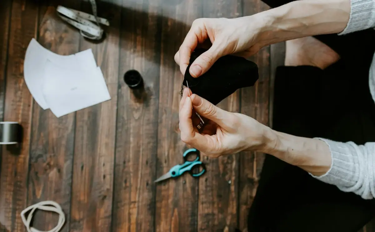 Person sewing black fabric with needle and thread, sewing kit, scissors, measuring tape, face masks, and thread on a wooden table.