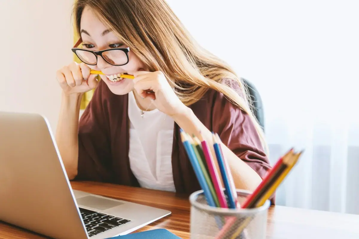 A woman with glasses biting a yellow pencil while working at her desk, with colored pencils in a cup nearby and an open laptop in front of her.