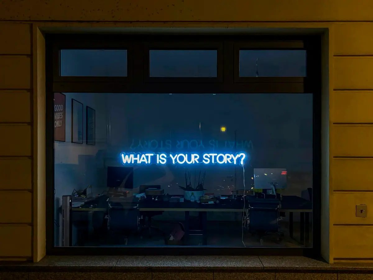 A storefront window with a neon sign that reads 'WHAT IS YOUR STORY?' inside an office with chairs, desks, and posters on the wall.