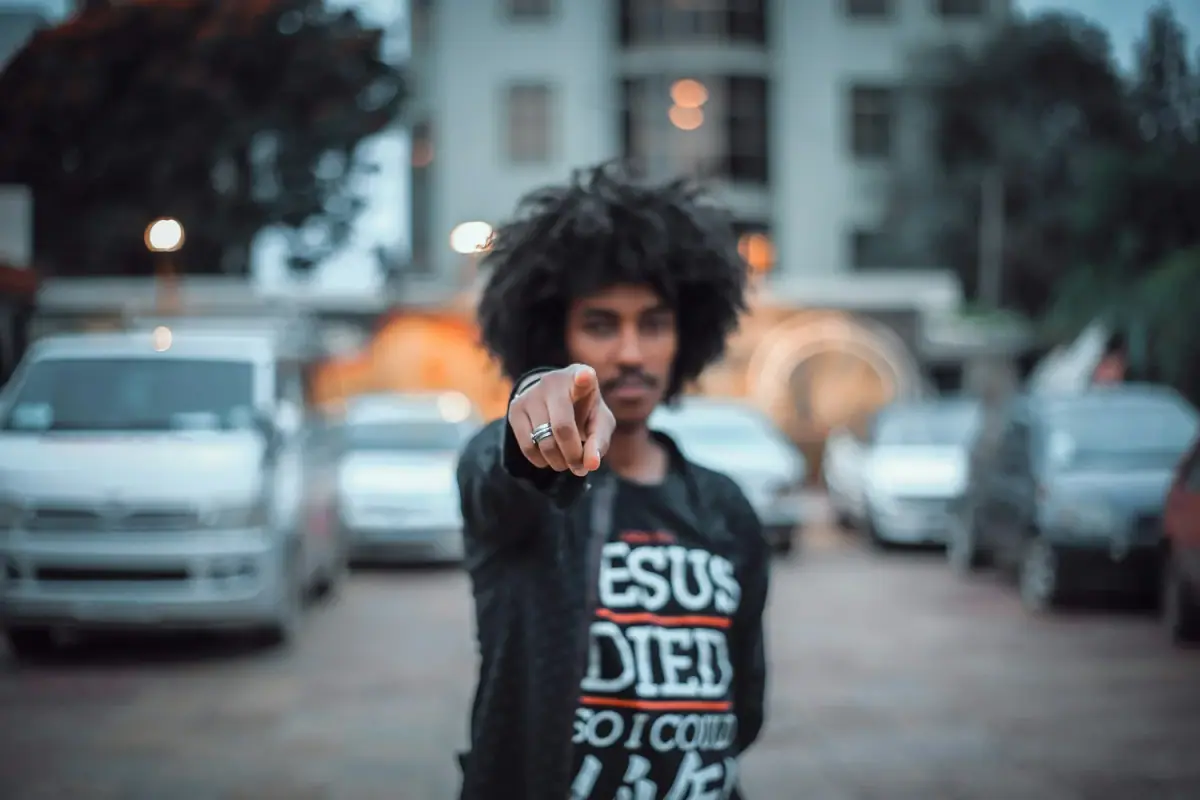 A person with curly hair pointing towards the camera with a parking lot and buildings in the background, blurry
