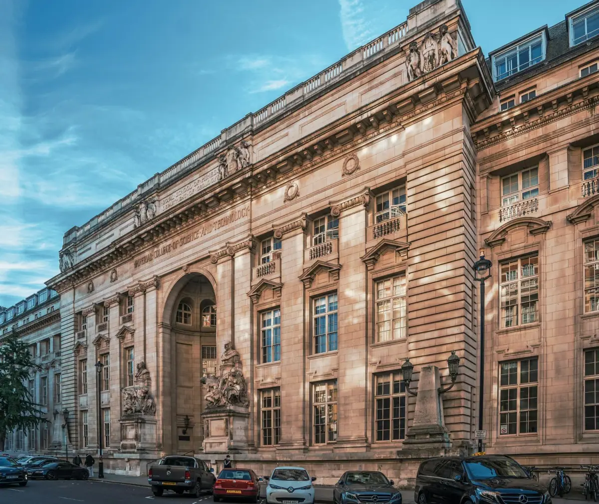 Historic building with classical architecture, large arched entrance, statues, and numerous tall windows, with cars parked in front and a tree on the left, under a blue sky.