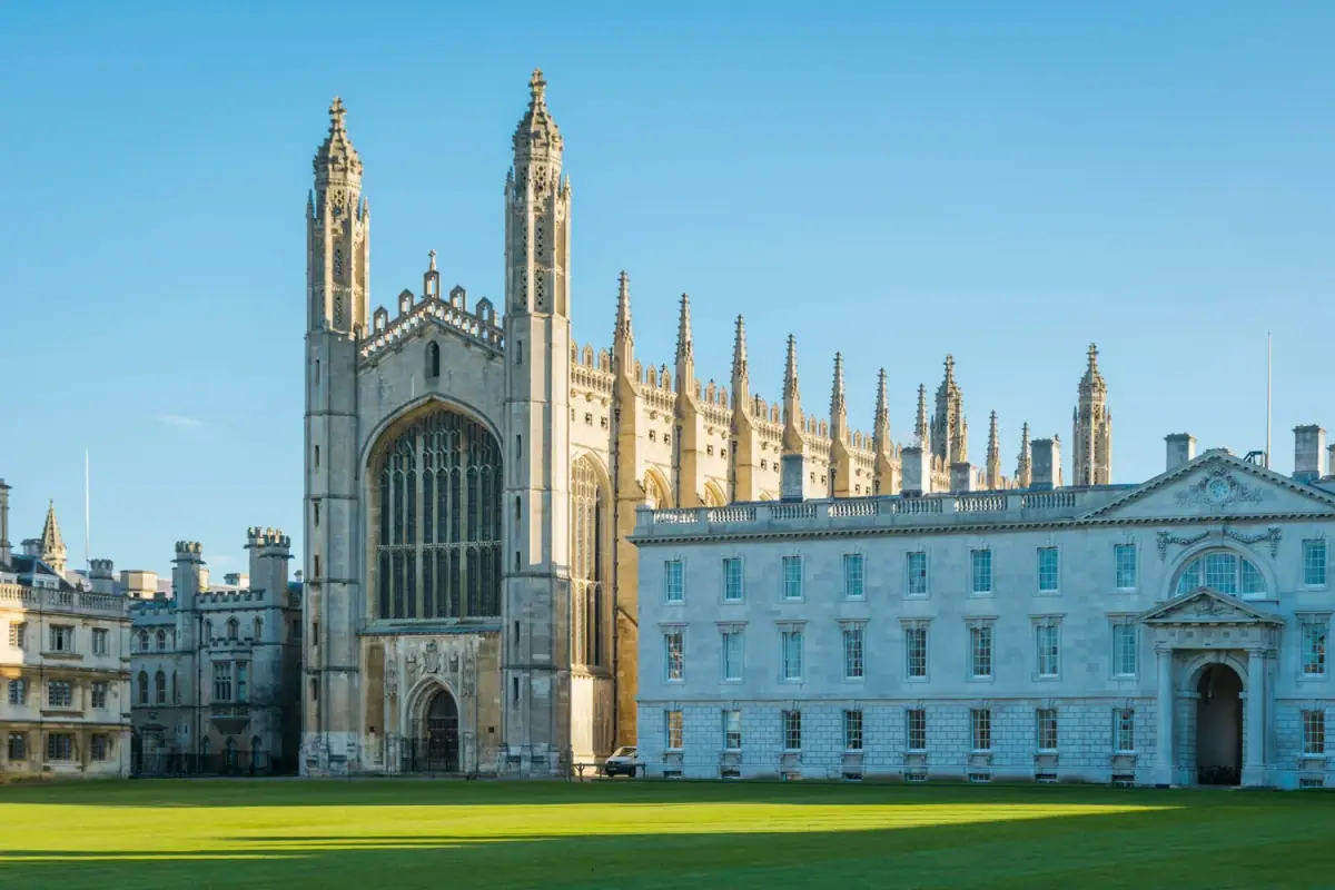A large historic Gothic-style cathedral with tall spires and intricate architecture, next to a neoclassical building, with a well-manicured lawn in the foreground under a clear blue sky.