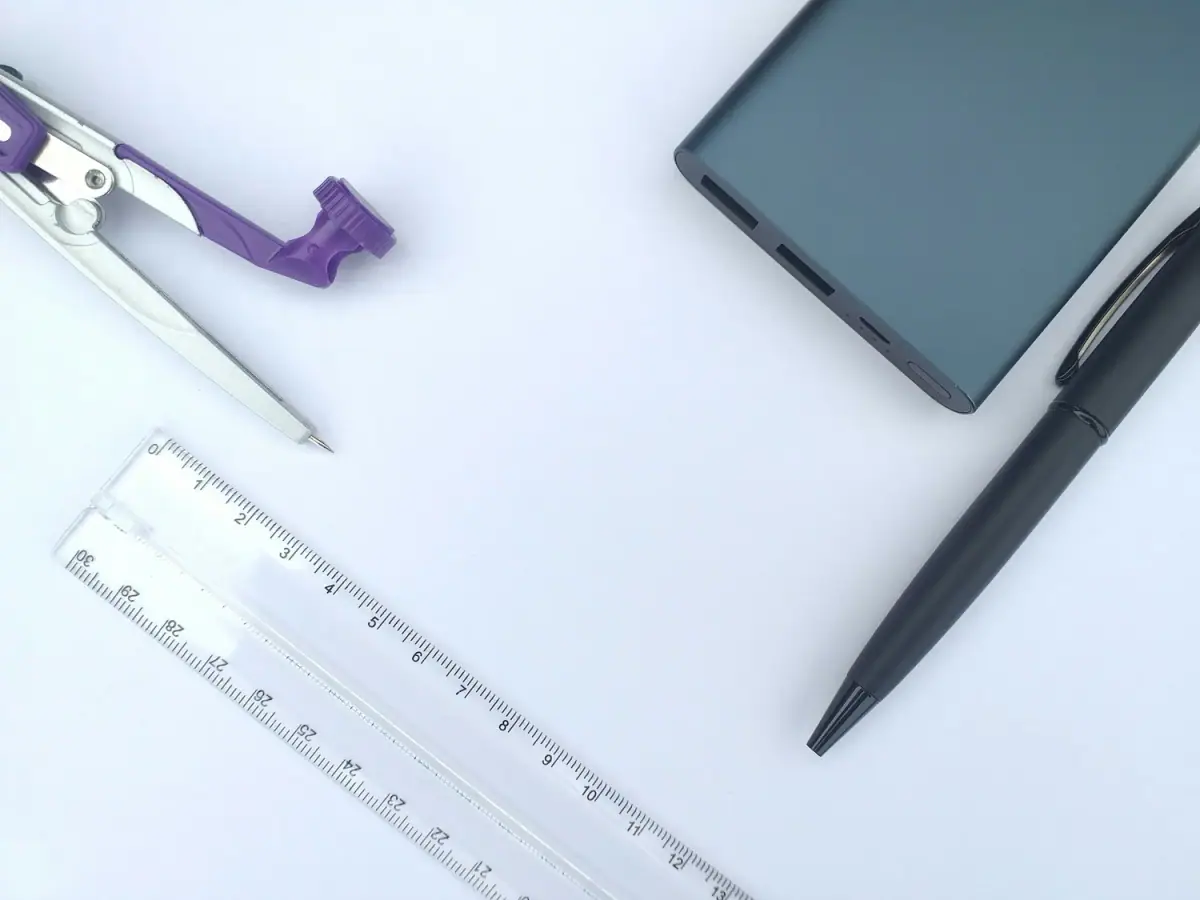 Office desk with purple and silver compass, clear ruler, gray external hard drive, black fountain pen, and turquoise laptop on a white surface.