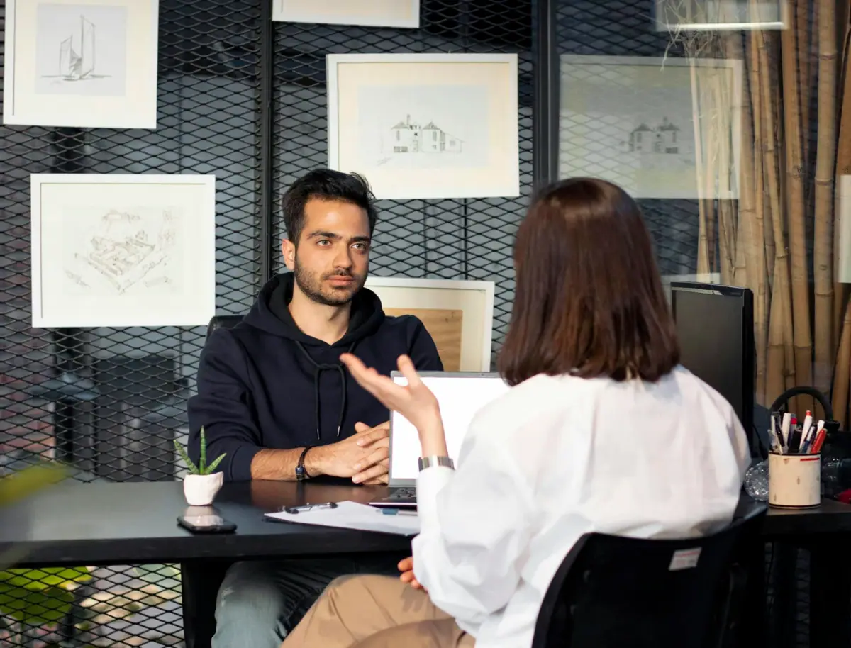 A man and a woman sit across from each other at a black table engaged in conversation in an office. The woman is gesturing with her hand. The background features framed architectural drawings and large windows with curtains.