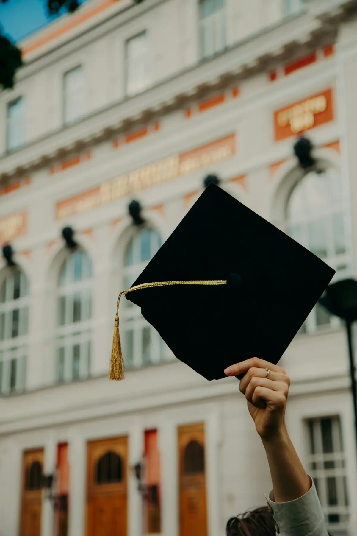 Person holding up a black graduation cap with a gold tassel in front of a historic building with large windows and decorative architectural details.