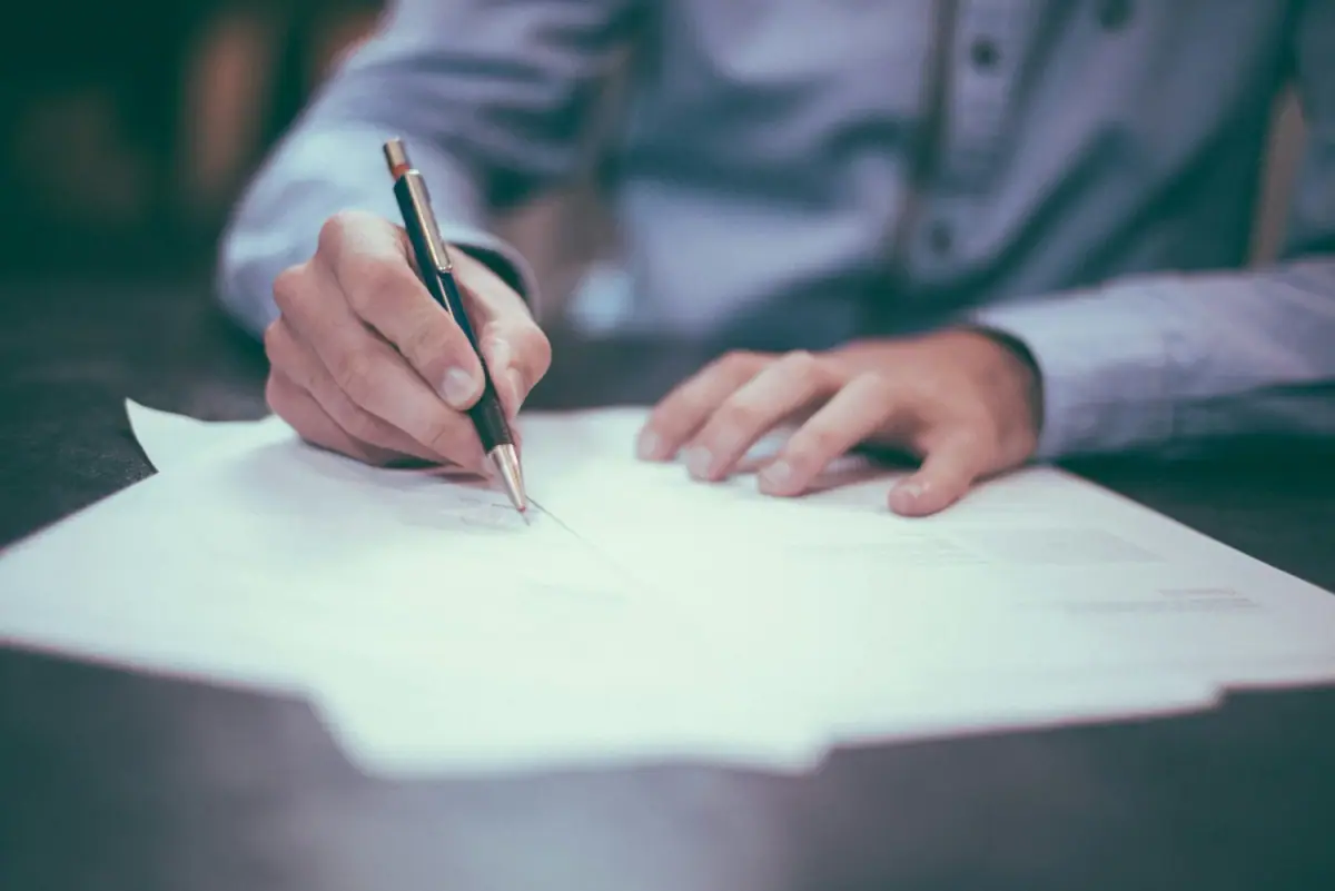 A person in a striped shirt is signing a document with a black and gold pen on a dark desk.