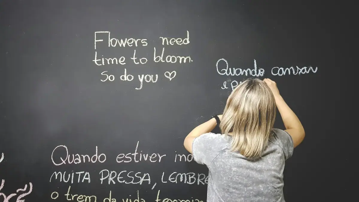 A girl writing on a blackboard with colorful chalk. The blackboard contains text in Portuguese and English about flowers, blooming, and life.
