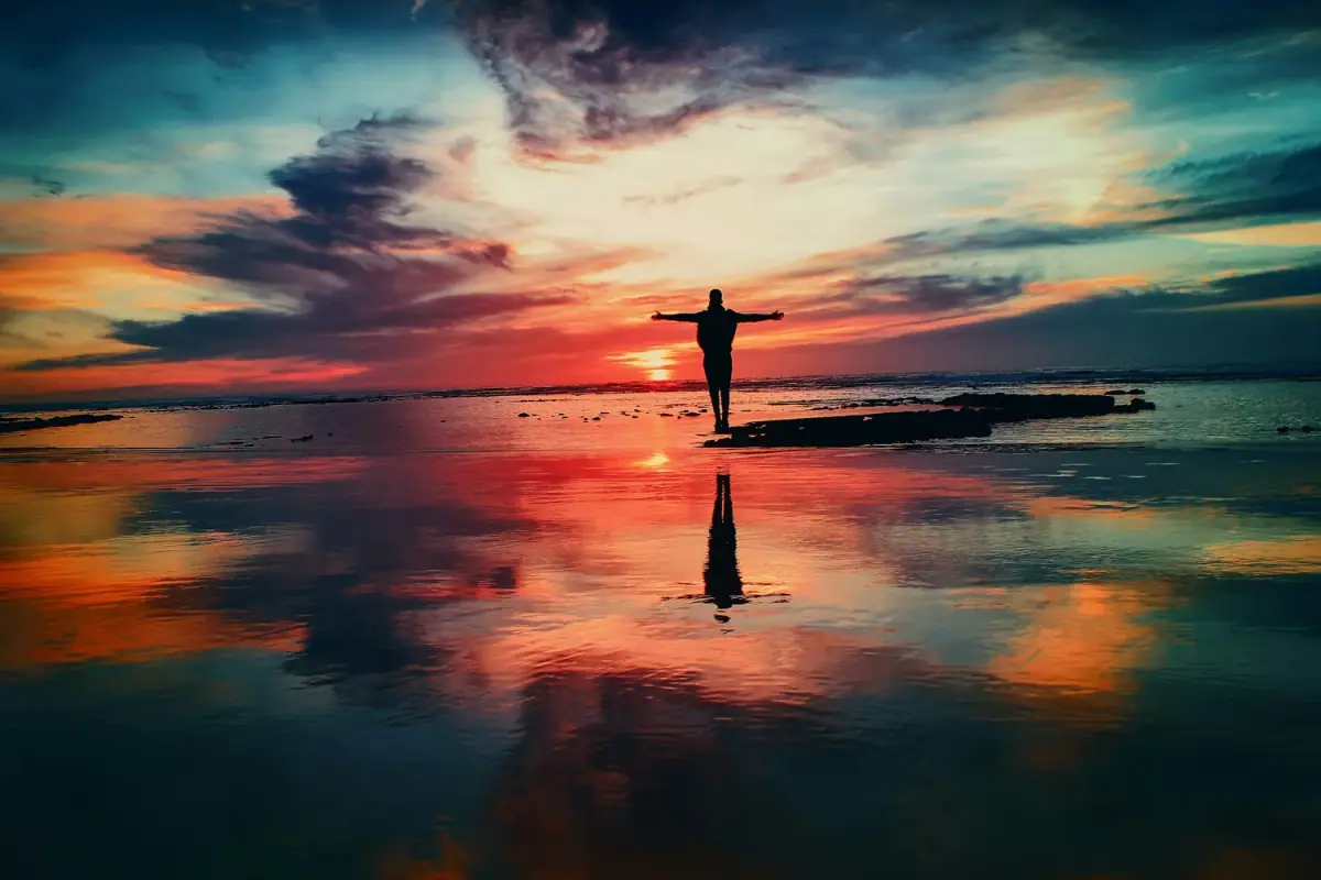 Person standing on a rock at the beach during sunset with arms outstretched and colorful sky with clouds reflected in the water.