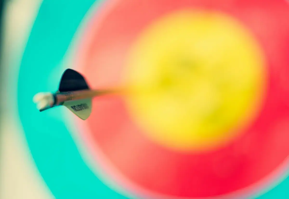 Close-up of a dart hitting the bullseye on a colorful dartboard with a blurred background of blue, pink, and yellow.