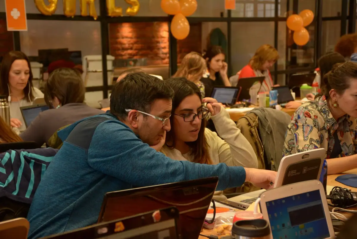 A tutor leaning in to help a student work through a problem on a laptop in a busy study session.