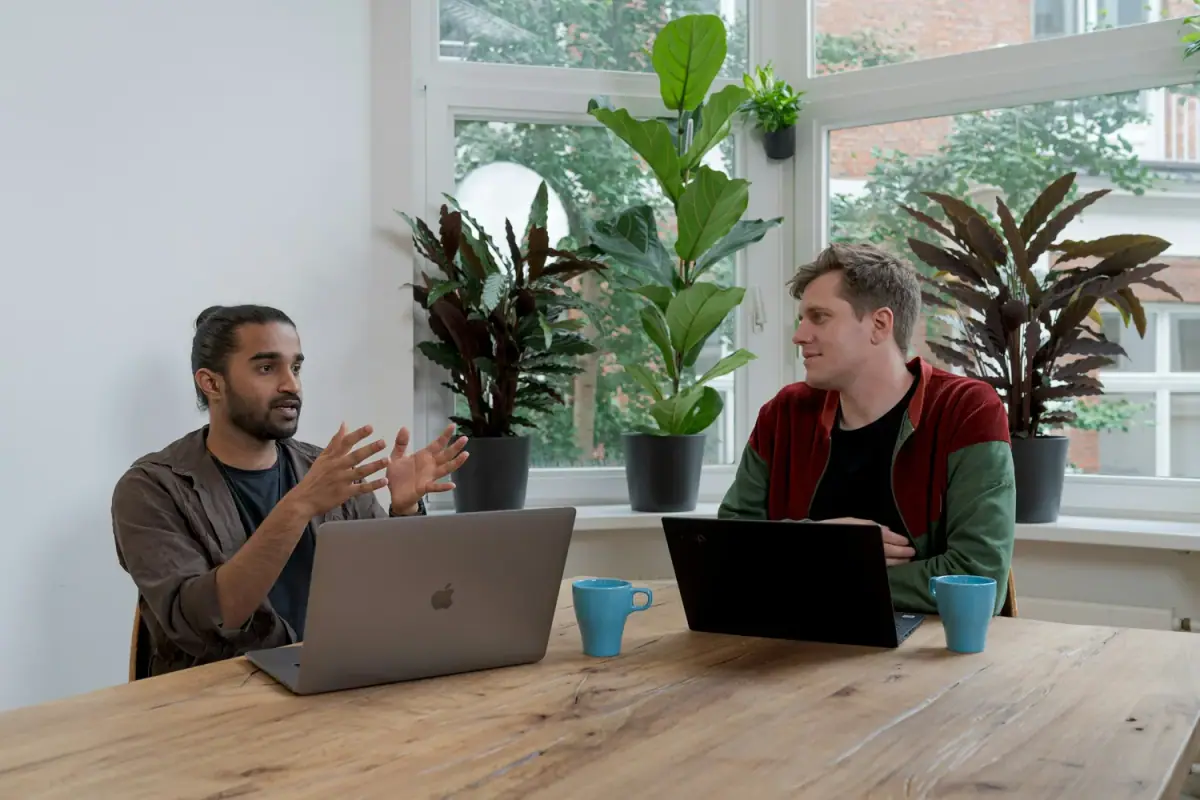Two men having a discussion in a bright room with large windows and plants, sitting at a wooden table with laptops and blue mugs.