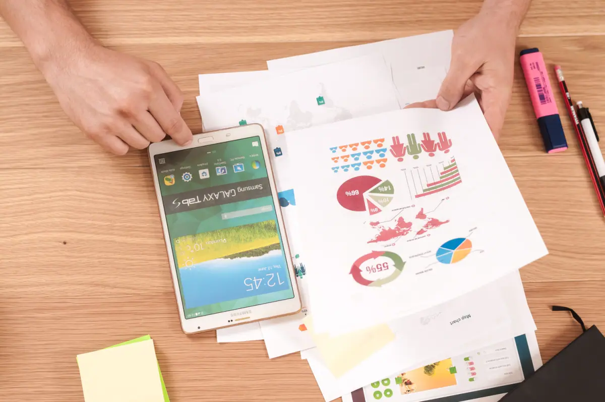 Person organizing printed business charts and graphs on a wooden desk, with a Samsung Galaxy Tab 4 tablet, pink and black pens, and sticky notes nearby.