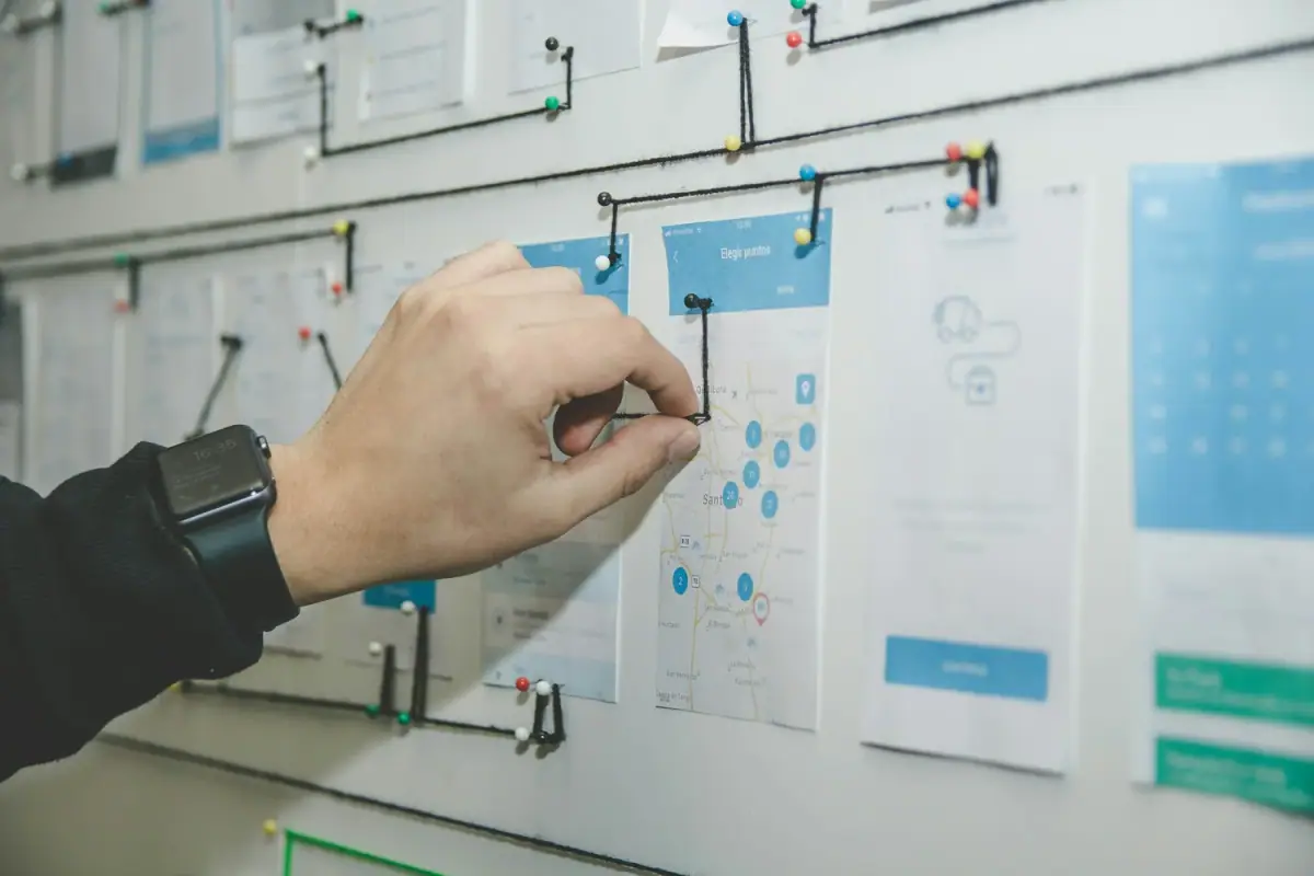 A person is pinning a paper map with blue stickers and black pins on a whiteboard with black strings and colorful push pins.