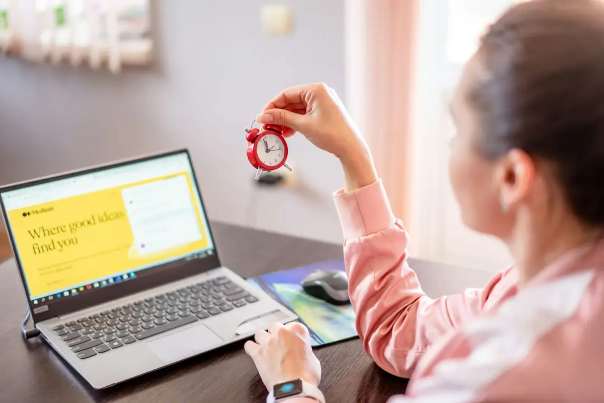 A woman sitting at a desk holding a small red alarm clock in her right hand, with a laptop open in front of her displaying yellow and white content, and a computer mouse on a mouse pad beside the laptop.