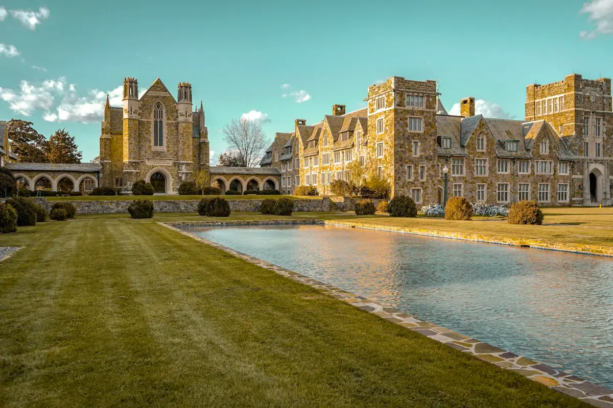 Large stone castle-like building with a reflection pool in front, surrounded by manicured lawns and bushes under a partly cloudy sky.