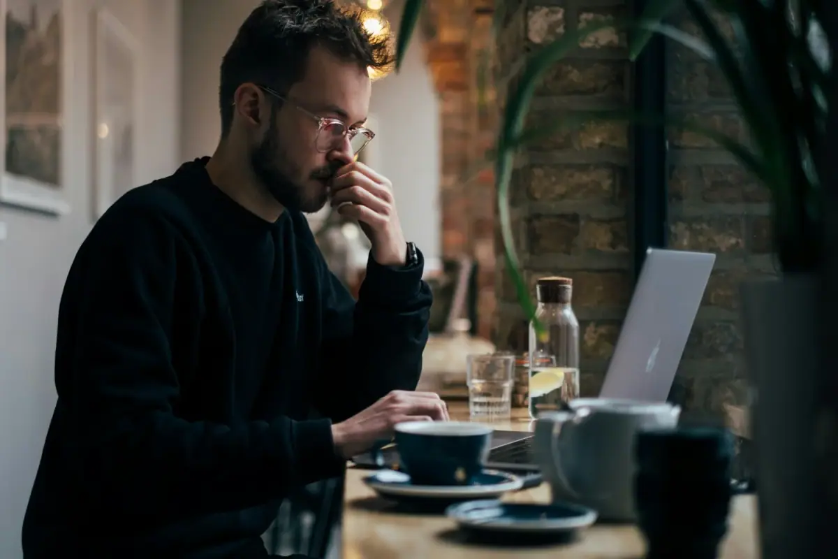 A young man with glasses and a beard sitting at a cafe table looking at a laptop, with a glass of water, cups, and a plant nearby.