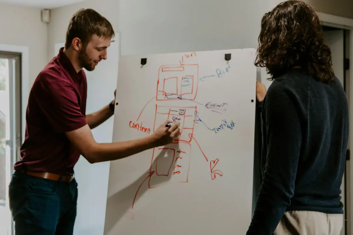 Two people discussing a whiteboard filled with sketches and notes about a website or app design, including sections labeled 'content', 'bio', and 'event'.