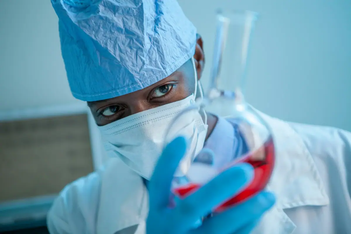 A scientist wearing a protective surgical mask, gloves, and a cap, holding a test tube filled with red liquid.