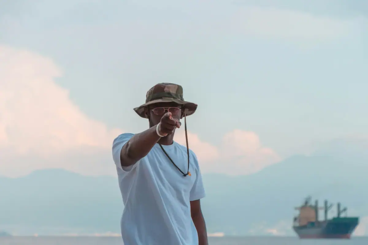 A man wearing a camouflage bucket hat, pink sunglasses, and a white t-shirt points towards the camera, with a large cargo ship sailing in the background on the water under a cloudy sky.