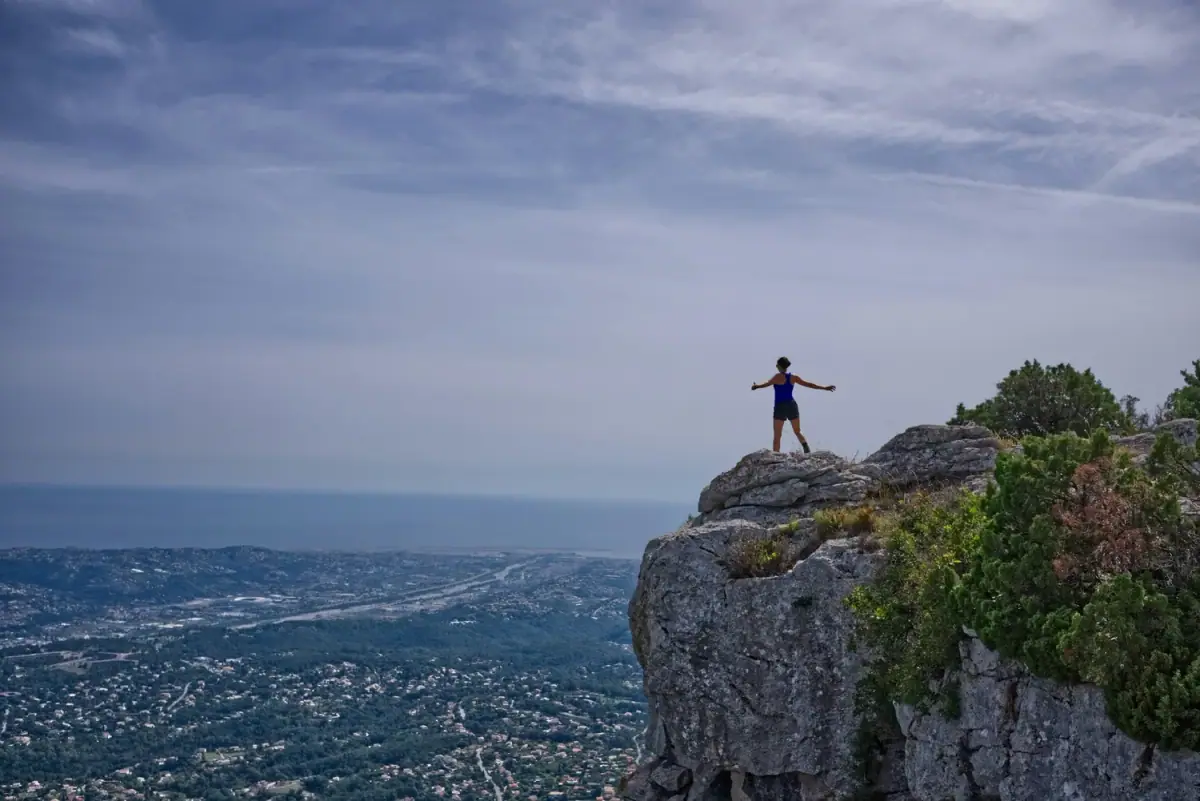 A person standing on a rocky cliff with arms outstretched, overlooking a cityscape and body of water under a partly cloudy sky.