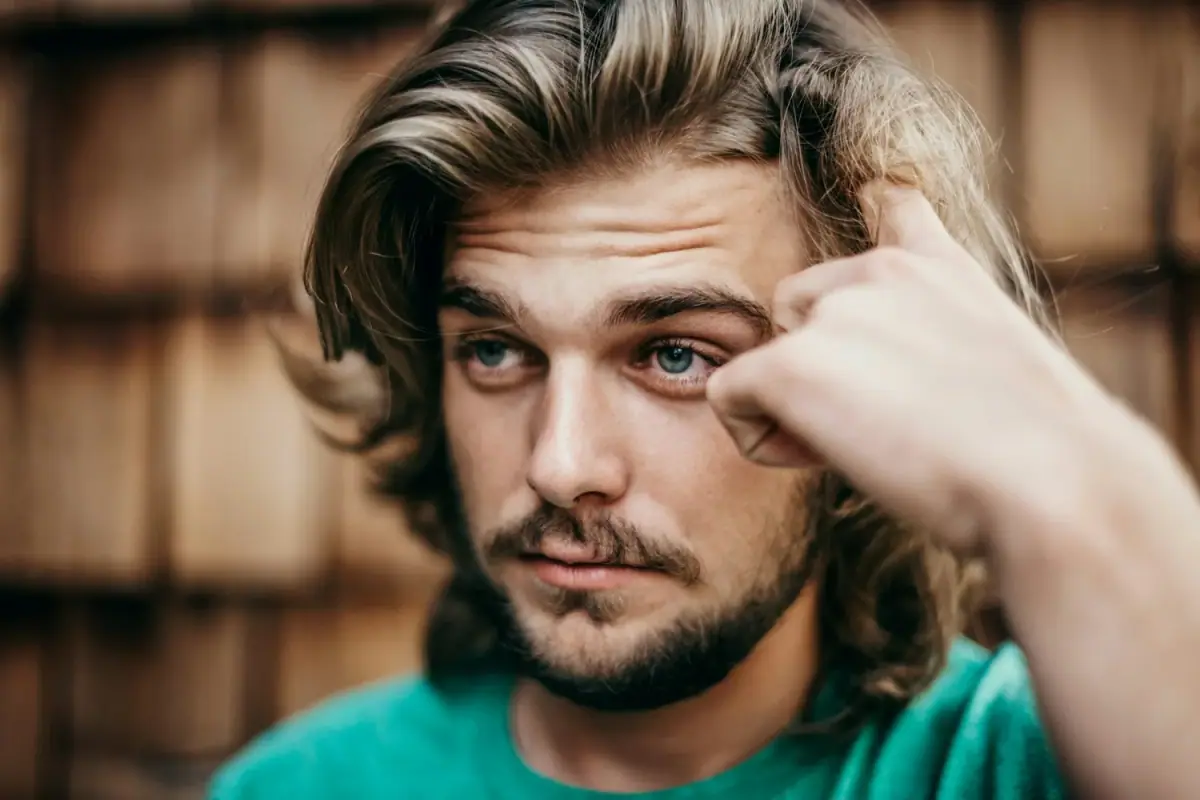 A young man with long hair pushing his fringe back from his forehead, looking thoughtfully towards the camera.