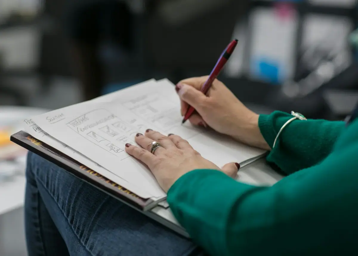 Person with painted nails and a ring on their finger writing in a notebook with a red pen, sitting with papers on their lap, in a casual setting.