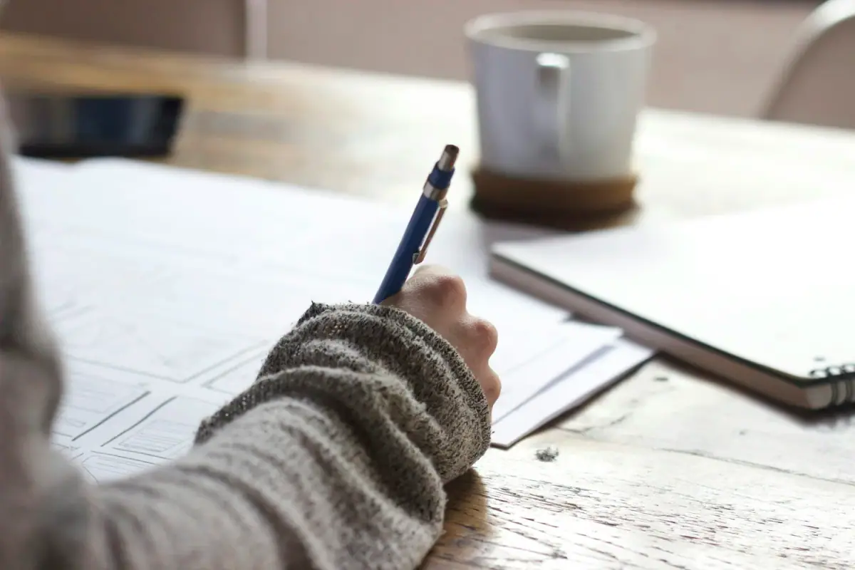 Person writing on a sheet of paper with a blue pen, other papers and a notebook on a wooden table, a white mug in the background