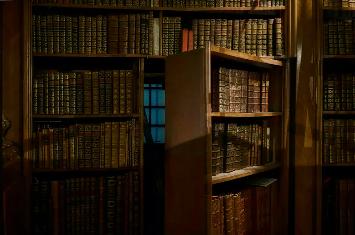 Wooden bookshelves filled with old, leather-bound books in a dimly lit library.