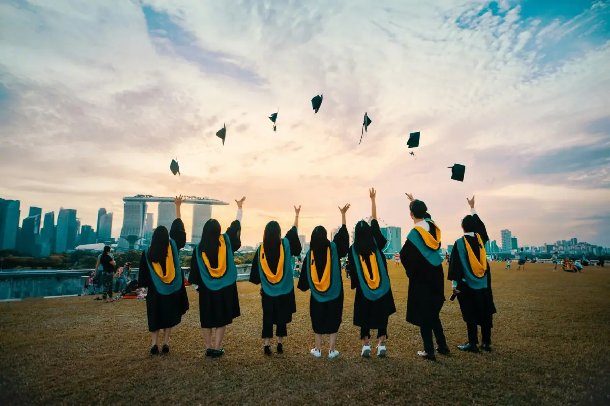 Group of graduates in caps and gowns celebrating outdoors with city skyline in the background, tossing caps into the air at sunset.