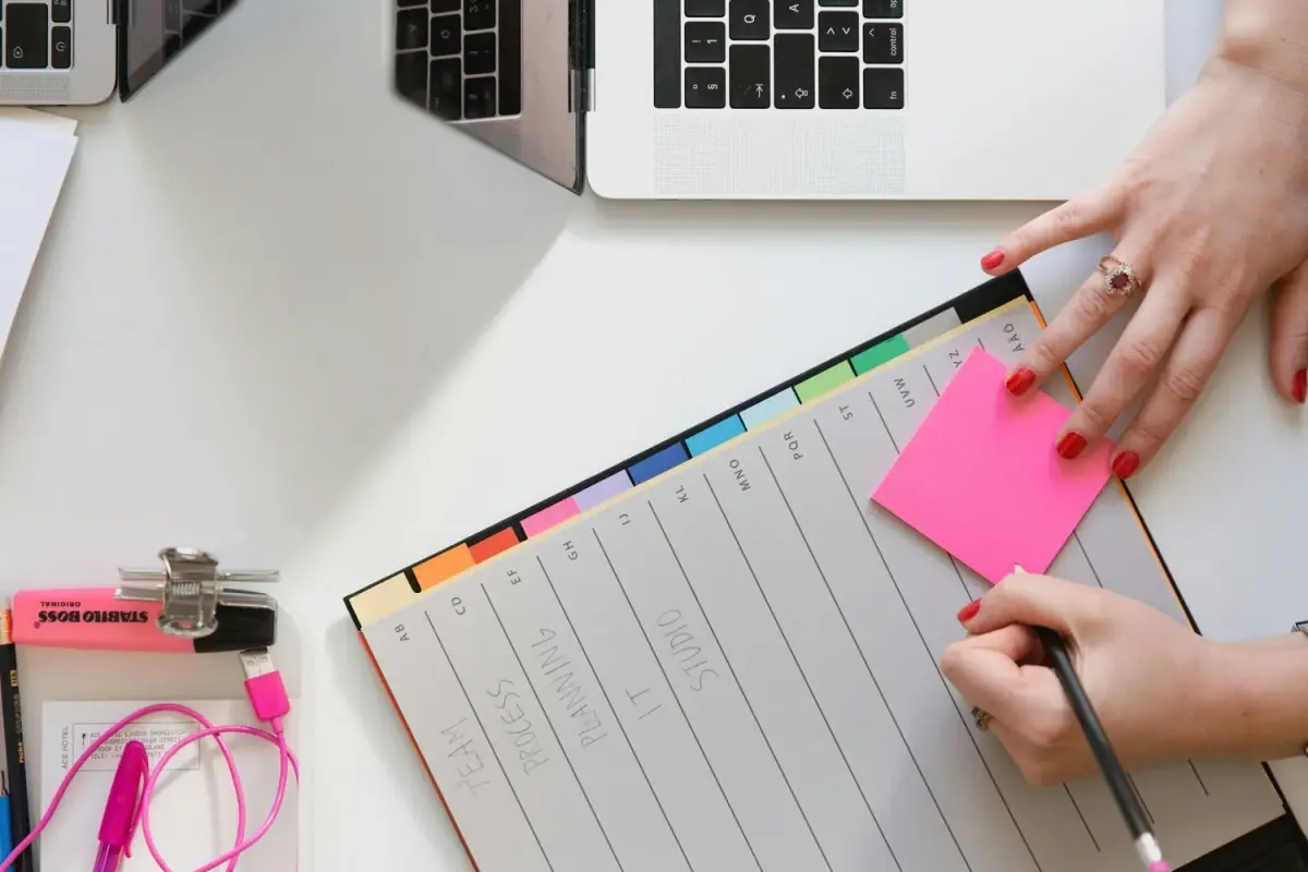 A person's hand with red painted nails writing on a pink sticky note, with a weekly planner visible on the table, along with a laptop, pink earphones, and other office supplies.