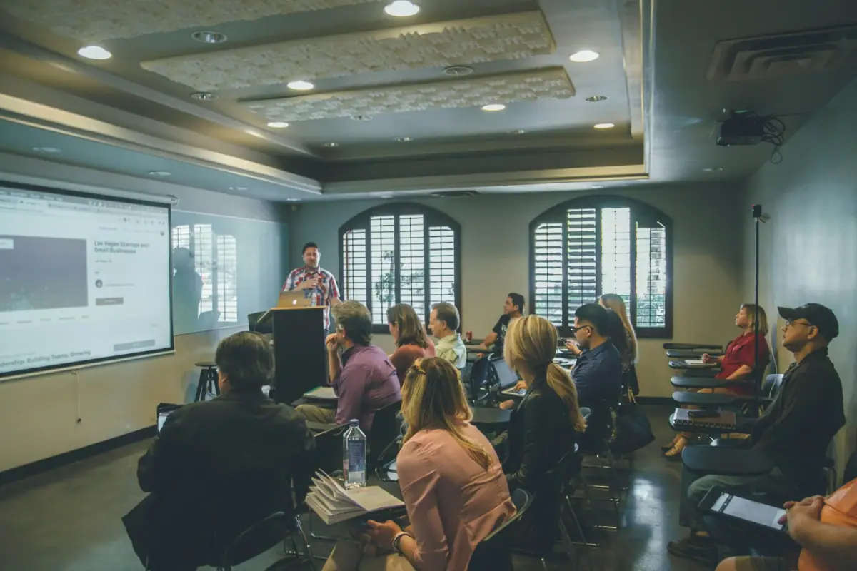 A man giving a presentation to a seated audience in a conference room with large windows and a projector screen.