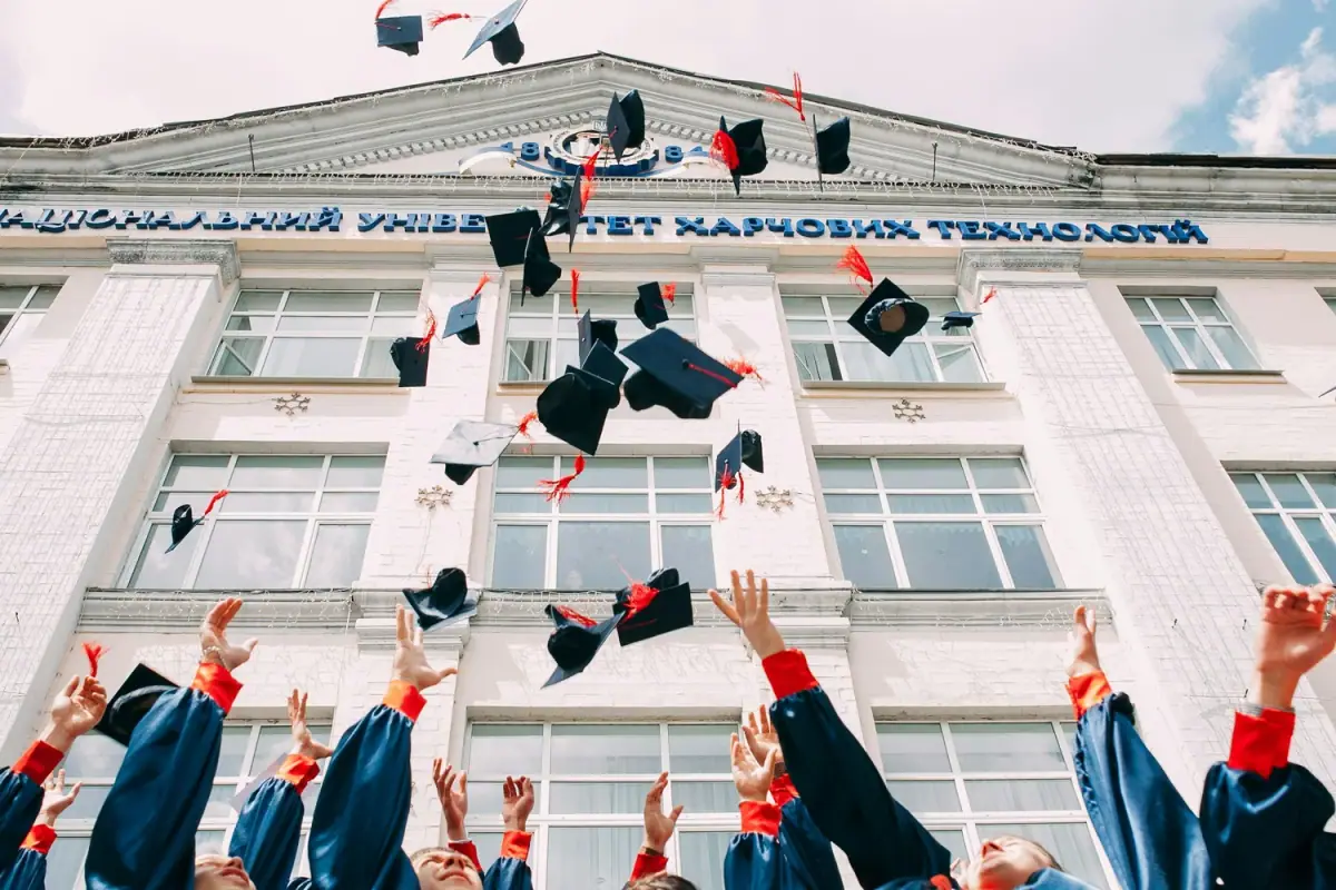 Graduates in gowns throwing caps into the air outside a university building with Ukrainian Cyrillic writing on the facade.